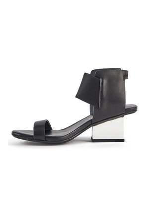 Sandal negro de punta abierta con una amplia correa en el tobillo y un bajo tacón de bloque plateado reflectante sobre fondo blanco.