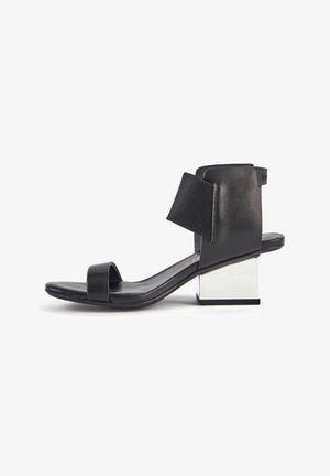 Sandale noire à bout ouvert avec un large strap à la cheville et un bas bloc argenté réfléchissant sur fond blanc.