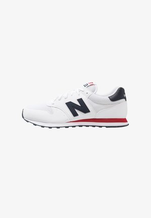 Zapatilla deportiva blanca con material de malla y sintético, acentos en negro y rojo, prominente logo "N" y una suela de goma texturizada.