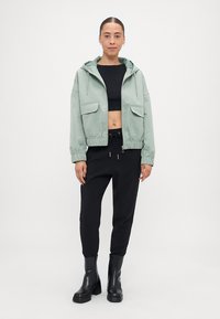 Veste cropped vert clair avec une capuche, deux poches avant, des poignets élastiques, portée par-dessus un crop top noir, associée à un pantalon noir et des bottes.