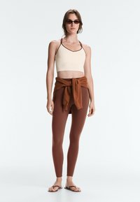 Femme portant des leggings marron, un crop top beige, un sweat marron noué à la taille, des lunettes de soleil et des sandales marron, debout sur fond blanc.