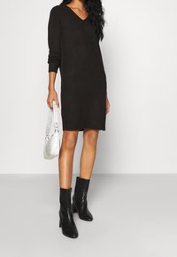 Robe-pull en tricot marron avec un col en V, des manches longues et une coupe légèrement ample. Associée à des bottines noires et un sac à main texturé blanc.