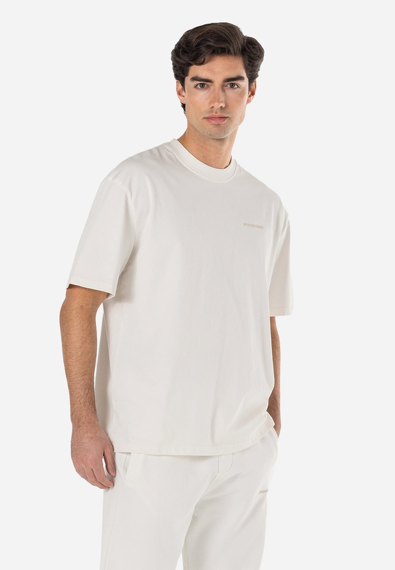 Camiseta de algodón de color beige claro con mangas cortas, cuello redondo y un sutil estampado de logo en el pecho. Textura suave y corte relajado.