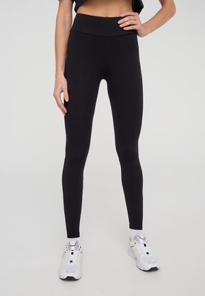 Leggings neri a vita alta realizzati in tessuto elastico, design aderente, superficie liscia e senza motivi visibili. Indossati con scarpe da ginnastica bianche.