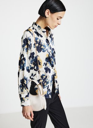 Blusa floreale con fiori blu e oro su sfondo bianco, stile con bottoni. Borsa chiara abbinata con tracolla a catena.