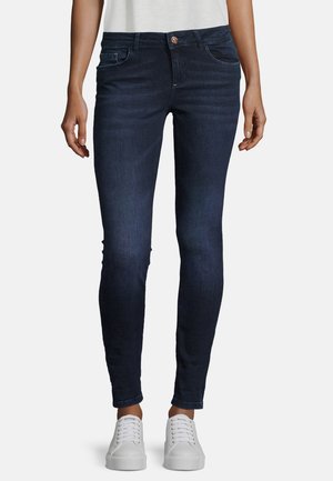 Jean slim bleu foncé porté par une personne, associé à des baskets blanches, visibles de la taille aux pieds sur un fond uni.