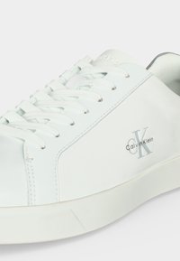 Λευκό χαμηλό παπούτσι Calvin Klein με λευκά κορδόνια και λογότυπο τυπωμένο στο πλάι, εμφανιζόμενο σε κοντινή λήψη σε μονόχρωμο φόντο.