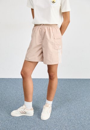 Personne portant un short beige Adidas avec des poches latérales, des baskets blanches Adidas et un t-shirt blanc, debout sur un sol recouvert d’un tapis bleu.