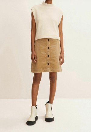 Personne portant un haut en maille blanc sans manches, une jupe en velours côtelé beige avec des boutons à l'avant et des bottines blanches avec des semelles noires.