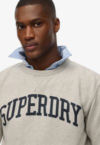 Lichtgrijze sweatshirt met "SUPERDRY" in gedurfde marineblauwe letters. Draag het over een blauw overhemd met kraag, waarbij de textuur van de kraag wordt benadrukt.