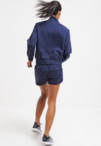 Chaqueta bomber de la Marina con tela texturizada y puños de canalé, combinada con shorts a juego. Bolsillo con cremallera visible en los shorts, se llevan zapatos deportivos.
