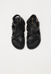 Black strappy sandals with a flat sole, featuring a cross-band design and an adjustable ankle strap with a gold buckle.