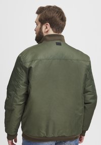 Maslinasto zelena bomber jakna s glatkom teksturom, rebrastim ovratnikom i manžetama, te malim crnim logotipom na stražnjem dijelu. Stražnji pogled.