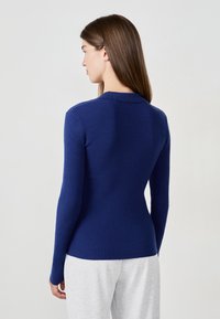 Kobieta z prostymi brązowymi włosami, ubrana w dopasowany granatowy sweter z długim rękawem i jasnoszare spodnie, zwrócona plecami do aparatu.