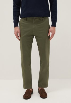 Man draagt olijfgroene slim-fit broek, donkerblauwe trui en bruine suède loafers, staand tegen een effen lichte achtergrond.