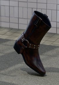 Σκούρο καφέ δερμάτινη μπότες μέχρι τον αστράγαλο με ασημένιες τρουκς και λεπτομέρεια με πόρπη, όρθια σε πλακόστρωτο δρόμο κοντά σε πλακάκια τοίχο.