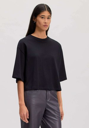 Femme aux longs cheveux foncés portant un haut noir ample à manches courtes et un pantalon en cuir foncé taille haute sur fond uni.