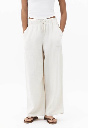 Persoon draagt roomkleurige wijde broek met trekkoortaille, wit tanktopje en bruine sandalen, handen in de zakken, staand tegen een witte achtergrond.