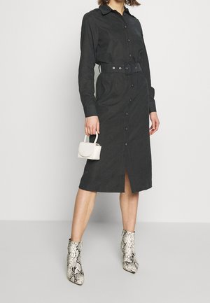 Vrouw die een zwarte midi hemdjurk met riem draagt, een kleine witte handtas vasthoudt, met enkellaarsjes met slangenprint tegen een effen achtergrond.