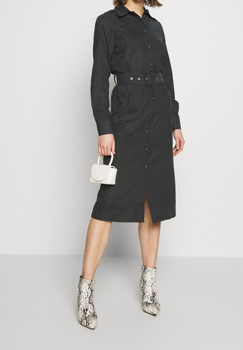 Femme portant une robe chemise midi noire ceinturée, tenant un petit sac à main blanc, avec des bottines à imprimé serpent sur un fond uni.