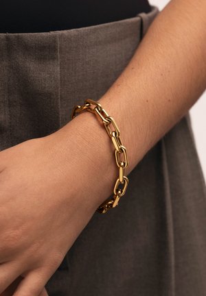 Pulsera de cadena de oro con eslabones alargados, acabado brillante, usada en una muñeca, sobre un fondo de tela gris.