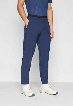 Mann trägt dunkelblaues Sportzeug mit schwarzem Bund und weißen Laufschuhen, steht mit einer Hand in der Tasche.