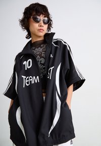 Černá sportovní bunda s bílými akcenty, s potiskem "TEAM 10". Krátké rukávy a zip; stylováno s černou vzorovanou košilí pod ní.