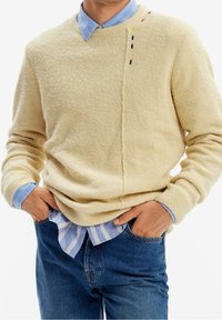 Pull en polaire beige avec une finition texturée, présentant un détail de couture et des accents de surpiqûres colorées. Associé à une chemise rayée bleu clair.