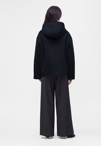 Personne aux longs cheveux vue de dos, portant une veste noire à capuche et un pantalon large et sombre sur un fond clair uni.
