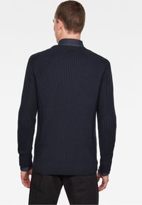 Ciemnoniebieski sweter prążkowany z wysokim kołnierzem i teksturowanym splotem. Posiada lekko opadające ramiona i jest zestawiony z dżinsową koszulą pod spodem.