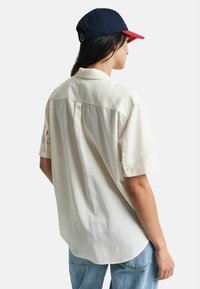 Cremefarvet kortærmet skjorte med knap-krave, bagfold og rundet søm. Bæres med lyseblå jeans og en marineblå og rød kasket.