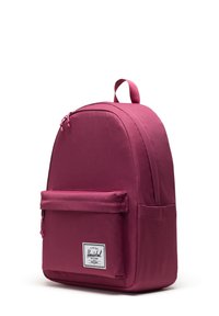Sac à dos en tissu mauve de forme arrondie, avec une poche avant zippée et une étiquette logo. Il est équipé de bretelles rembourrées et d'une poignée supérieure pour le transport.