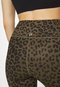 Olive zielone legginsy z czarnym nadrukiem w panterę. Zawierają płaskie szwy i małe białe logo na pasie. Elastyczny, dopasowany krój.