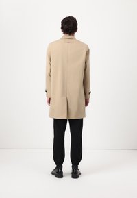 Beige trench coat with a tailored fit, long sleeves, back vent, and black button accents. Worn over black tapered pants and shoes.