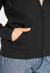 Haut à capuche zippé noir avec poches avant et poignets côtelés. Le tissu semble doux et texturé. Assorti avec un pantalon bleu clair.