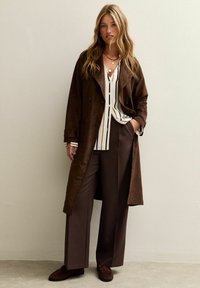 Abrigo largo de ante marrón sobre una camisa a rayas blanca y negra, combinado con pantalones anchos marrones y zapatos oscuros sin cordones, luciendo collares en capas.