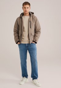 Chaqueta aislante beige con capucha forrada, bolsillo con cremallera en el pecho y detalles de botones a presión, llevada sobre un suéter ligero y unos vaqueros azules.