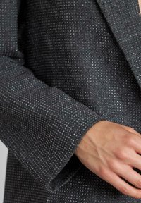 Grijze gestructureerde blazer met een houndstoothpatroon en subtiele zilveren accenten, met een gestructureerde revers en brede mouwen.