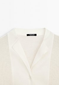 Chemisier blanc avec un col, présentant un design en tricot texturé, une fermeture à boutons et une étiquette portant la mention "Massimo Dutti". Tissu léger et aérien.