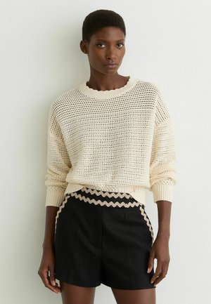 Kvinde med kort hår iført en cremefarvet åbenstrikket sweater og højtaljede sorte shorts med beige bølget kant, stående op ad en hvid væg.