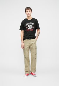 Tricou negru din bumbac cu text alb și roșu; pantaloni cargo khaki cu buzunare; adidași roșii. Ținută casual, cu un croi lejer.