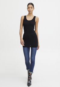 Schwarzes ärmelloses Tunika-Top kombiniert mit eng anliegenden blauen Jeans. Das Model trägt gemusterte Stiefeletten. Glatte Textur, lässiges Design.