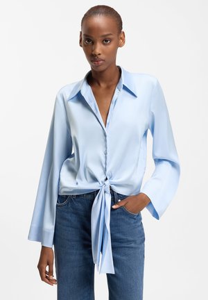 Woman wearing light blue long-sleeve blouse with large collar and front tie, paired with blue denim jeans, posing against white background.