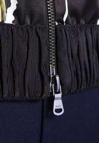 Veste noire zippée avec tissu plissé à l'ourlet, dotée d'un tire-zip argenté portant le logo "HUGO". Textile lisse avec des motifs contrastés.