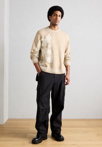 Νεαρός άνδρας φοράει ένα μπεζ πουλόβερ με λευκό σχέδιο καμηλοπάρδαλης, μαύρο παντελόνι cargo και μαύρα χοντρά μποτάκια, στέκεται σε ξύλινο πάτωμα.