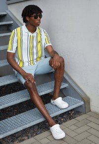 Polo bianco a maniche corte con strisce verticali gialle e nere, abbinato a pantaloni corti azzurri e ciabatte bianche, seduto su gradini di metallo.