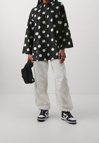 Chemise noire à gros pois blancs, manches longues, fermeture à boutons ; associée à un pantalon blanc ample et des baskets noir et blanc.