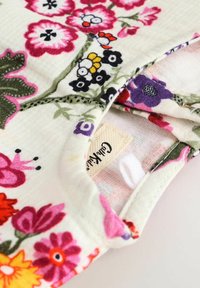 Tissu à motif floral avec des fleurs roses, violettes et noires sur un fond blanc doux ; comporte une étiquette cousue avec le nom de la marque.