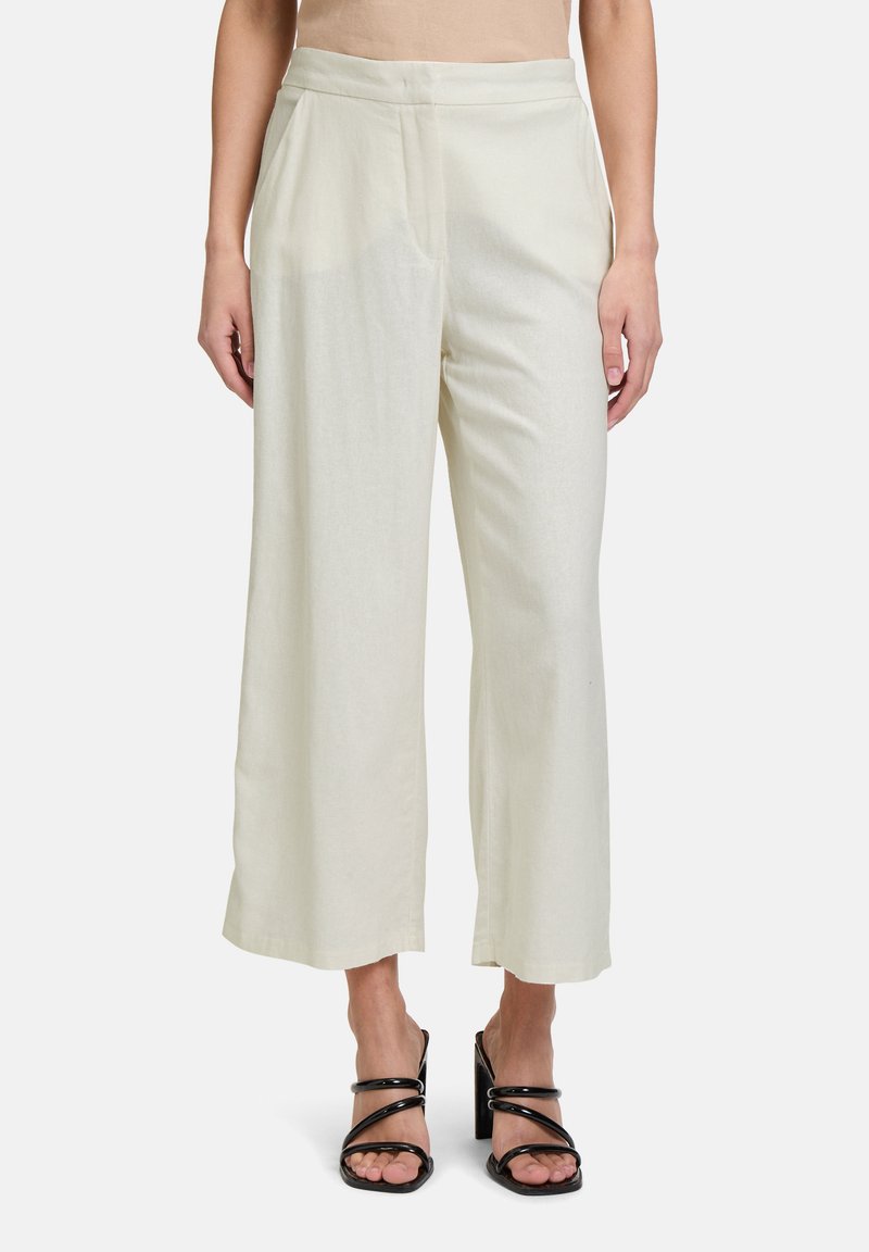 Wijdvallende broek van lichtcrème stof met een gladde textuur. Voorzien van een geperste plooi aan de voorkant en een aansluitende tailleband. Gepaard met zwart gespenhakken.