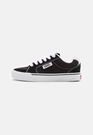 Zapatilla de lona negra con cordones blancos, suela de goma blanca, costuras en contraste, logo de Vans en el costado y un acento rojo en la parte trasera.